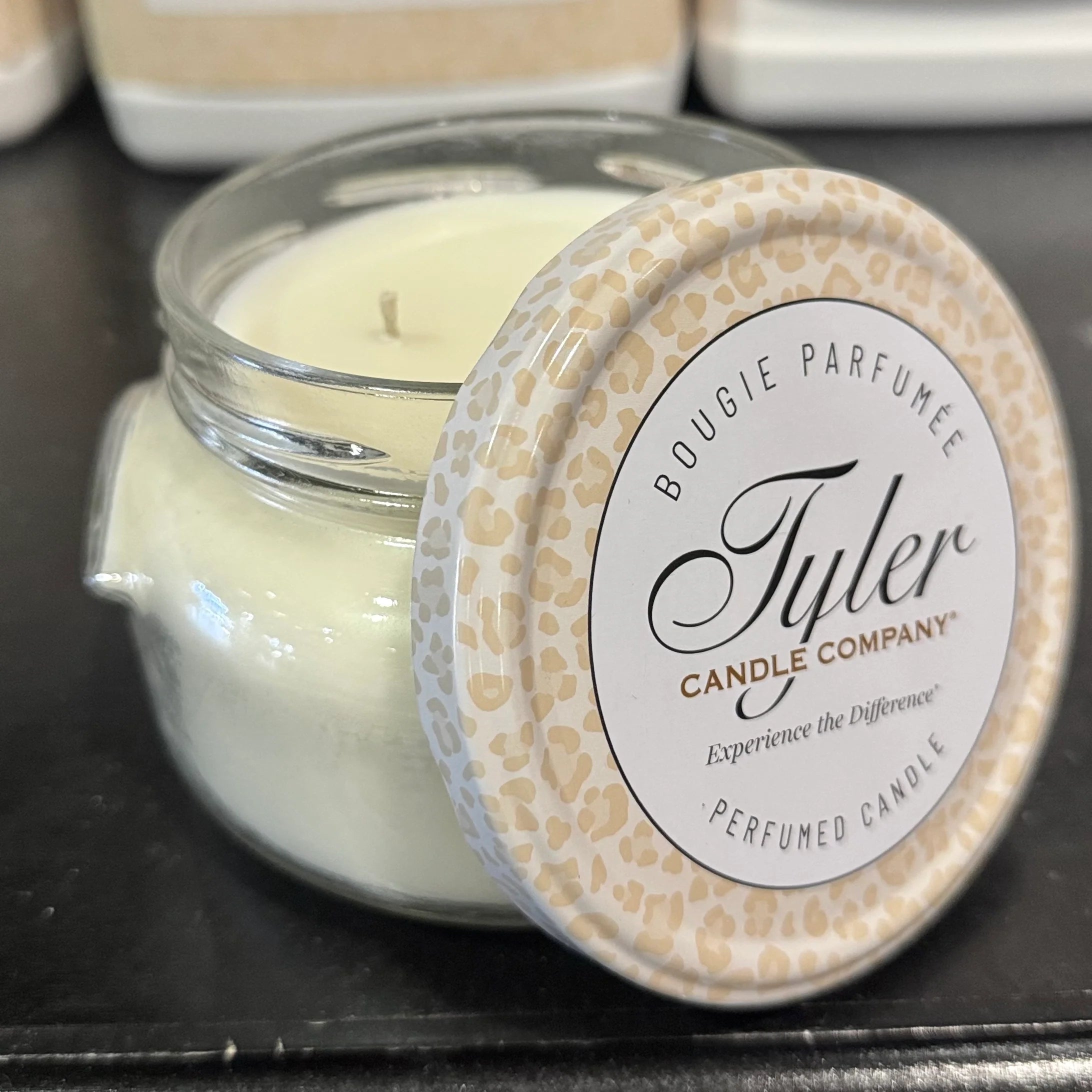 White candle in a glass jar with a decorative lid on a black surface, featuring the Tyler Candle Company branding.