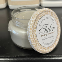 Silver candle in a glass jar with a decorative lid on a black surface, featuring the Tyler Candle Company branding.