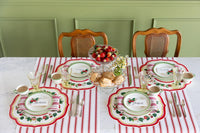 A styled holiday table setting with four matching place settings featuring red and white striped table linens, green and white plates with red berry motifs, silver cutlery, and a central fruit bowl on a white marble table against a green paneled wall.