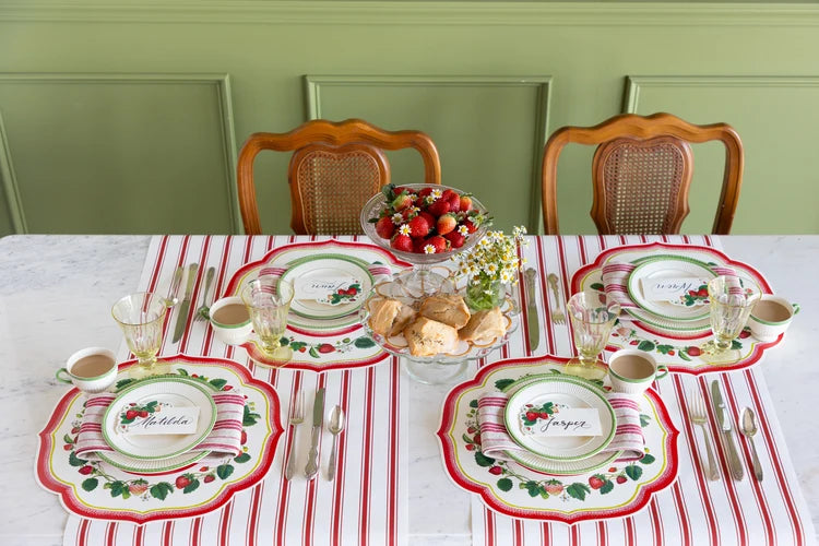 A styled holiday table setting with four matching place settings featuring red and white striped table linens, green and white plates with red berry motifs, silver cutlery, and a central fruit bowl on a white marble table against a green paneled wall.