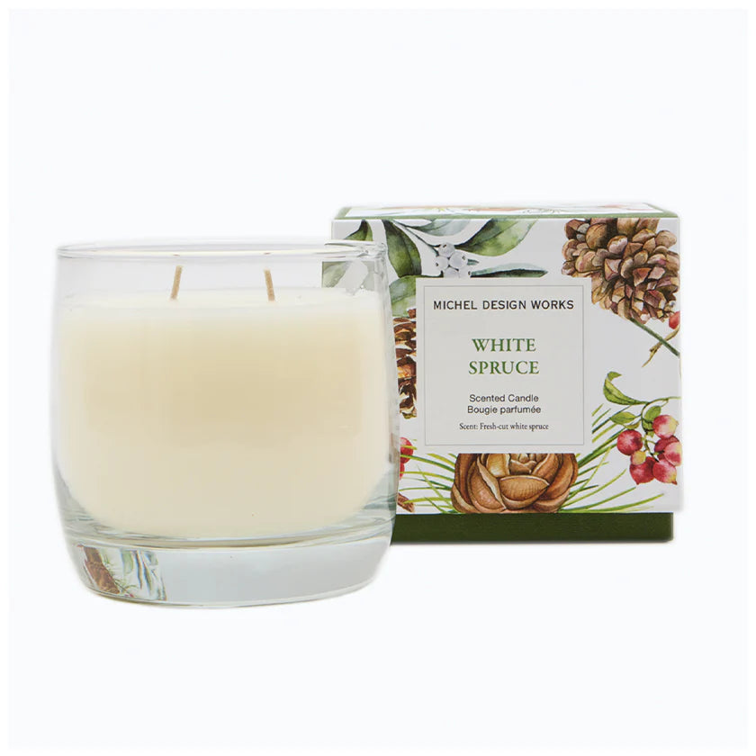 A Michel Design Works White Spruce scented candle in a clear glass jar with two wicks, shown next to its illustrated packaging box featuring pinecones and greenery.