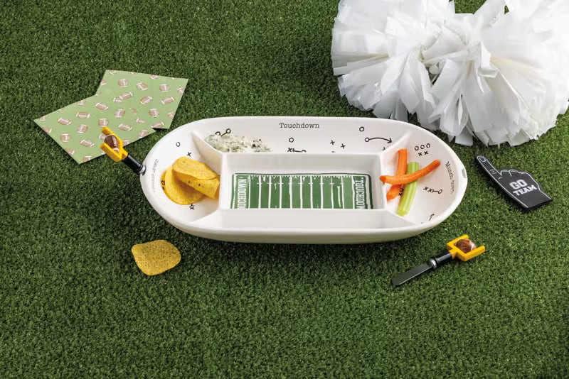 A football-themed divided serving platter set on artificial grass with snacks, napkins, a pom-pom, and a toy whistle.