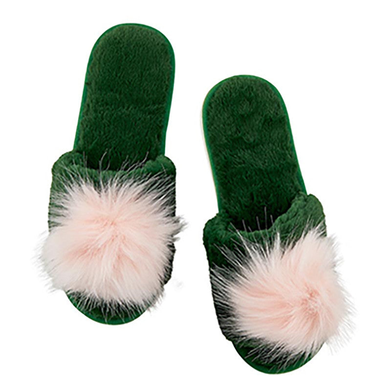 A pair of dark green plush slip-on slippers with large fluffy pale pink faux-fur pom-poms on the front, shown on a plain white background.