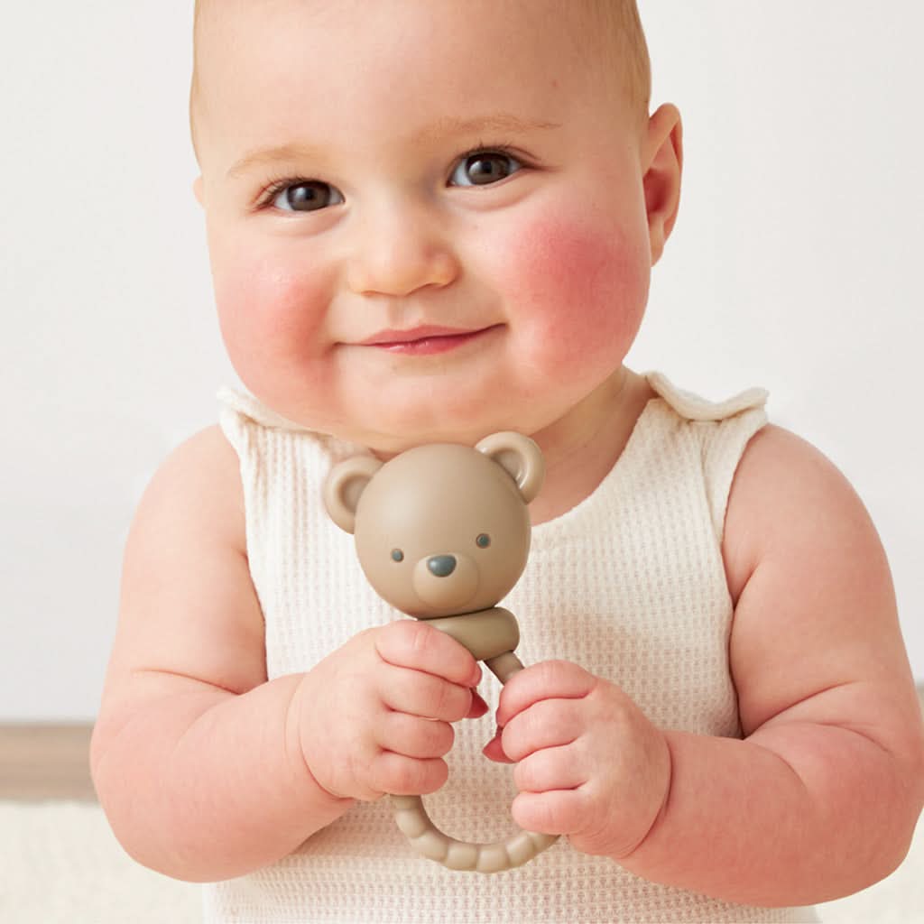 A beige silicone baby teether shaped like a teddy bear head with a textured ring handle, shown being held by a smiling baby.