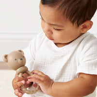 A beige silicone baby teether shaped like a teddy bear head with a textured ring handle, shown being held by a baby.