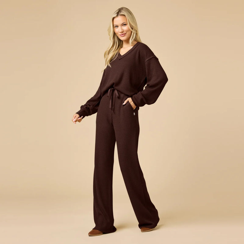 A woman modeling a dark brown knit lounge set consisting of a long-sleeve V-neck top with a tie waist and matching wide-leg pants, set against a solid beige studio background.
