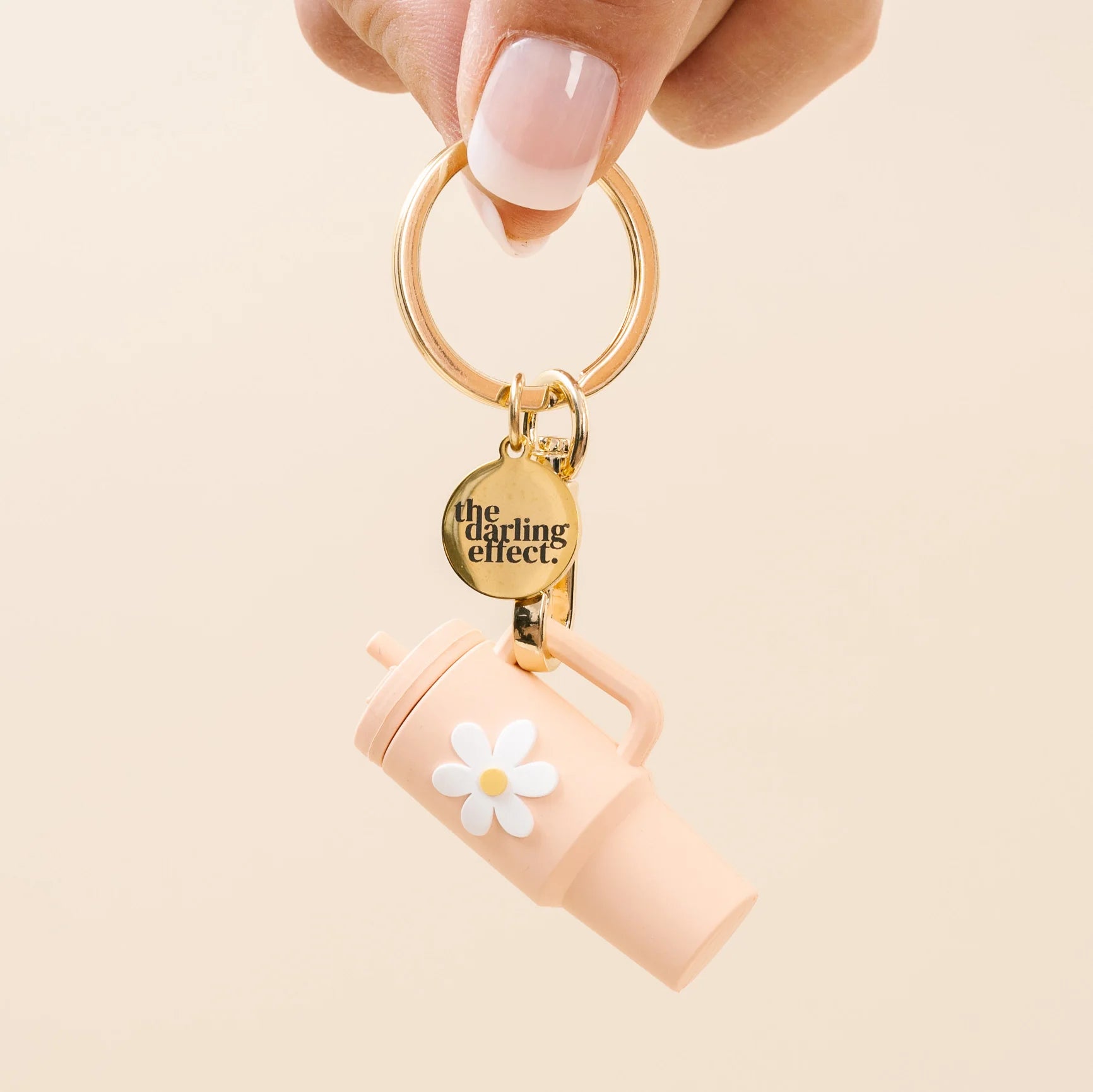 A hand with a French manicure holds a gold keyring featuring a light pink travel tumbler charm with a white daisy and a gold heart-shaped tag that reads 'the cycling effect.' on a solid pale beige background.
