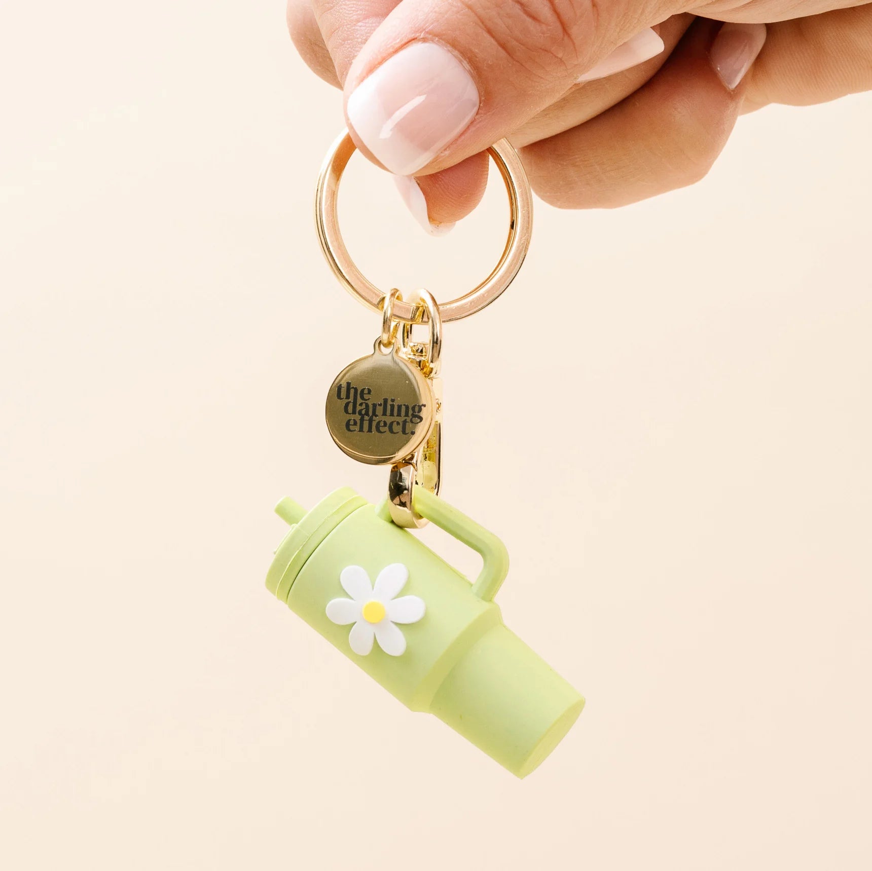 A hand with light pink manicured nails holds a gold keyring featuring a small light green tumbler charm with a white daisy flower and a round gold tag that reads 'the doting effect'.