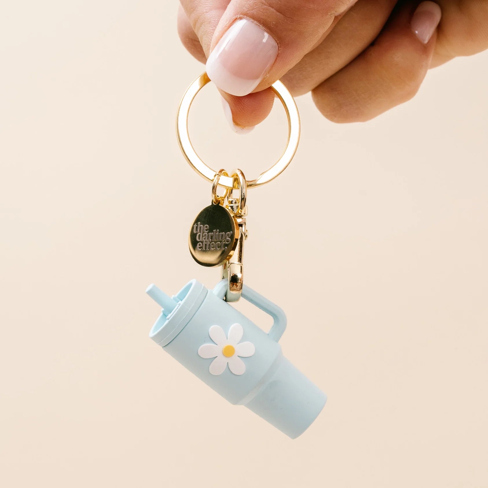 A hand holds a gold keychain featuring a light blue tumbler charm with a white daisy flower and a small round metal tag that reads 'the morning after'.