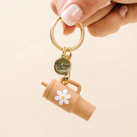 A hand with pale pink manicured nails holds a gold keyring featuring a small tan travel tumbler charm with a white daisy and a round gold tag that reads 'the darling effect'.