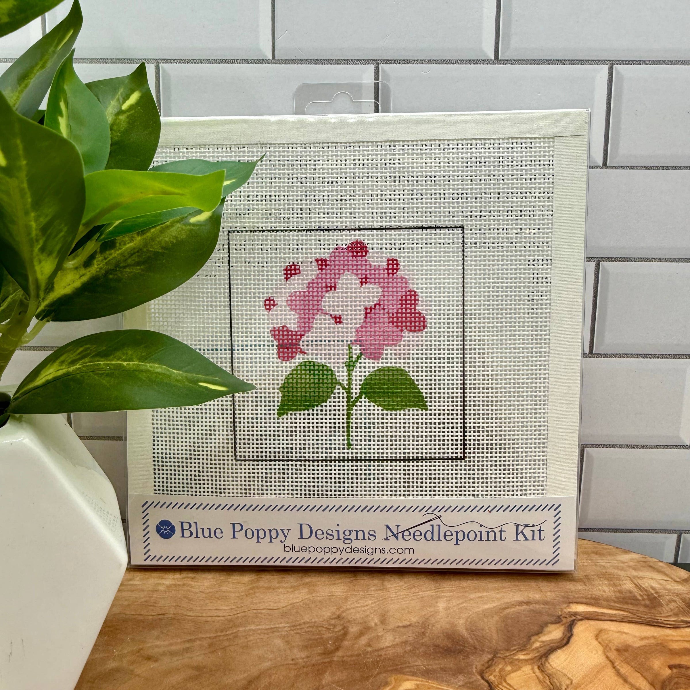 Cross-stitch kit with pink flower design on a wooden surface with a plant and tiled wall background.
