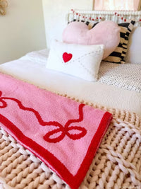 A pink knit blanket with a red border and bow design draped over a bed, alongside a white pillow with a red heart and other decorative pillows in a cozy bedroom setting.