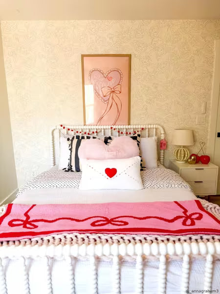 A styled bedroom scene featuring a white rope bed frame decorated with pink and white bedding, a pink throw blanket with red bow patterns, and a white pillow with a red heart. A pink framed art print hangs on the textured wallpapered wall above the headboard.