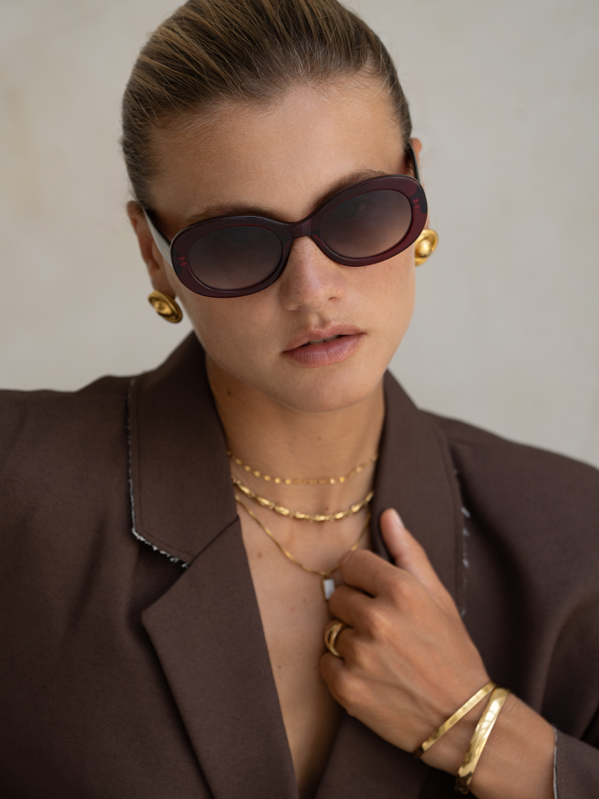 Woman wearing sunglasses, gold earrings, necklaces, and bracelets against a neutral background