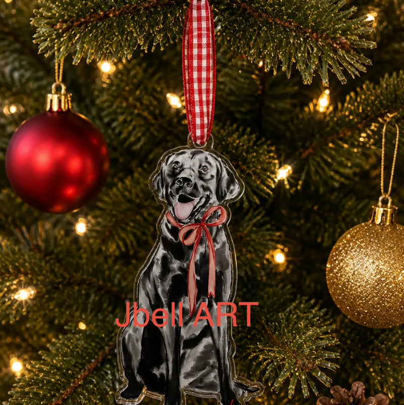 A custom Christmas ornament shaped like a black Labrador Retriever wearing a pink bow, hanging from a red and white gingham ribbon on a decorated Christmas tree. The ornament features a red and white gingham ribbon at the top and a pink bow on the dog's neck.