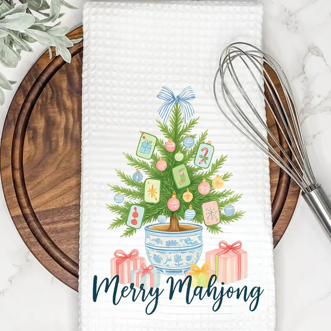 Decorative towel with a Christmas tree design and 'Merry Mahjong' text, placed on a kitchen surface with a whisk and cutting board.