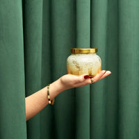 Hand holding a glass jar with a gold lid against a green curtain background