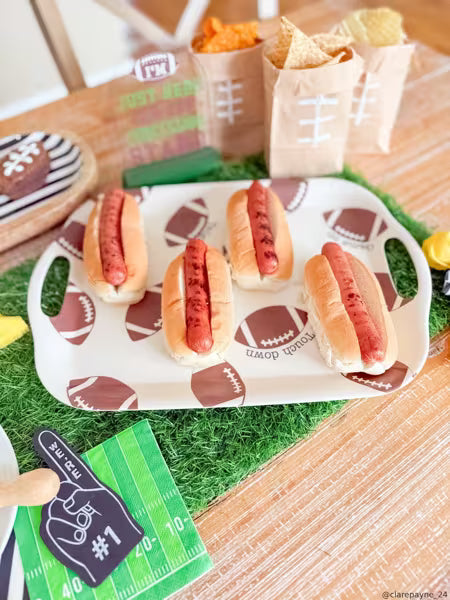 A football-themed hot dog platter with four hot dogs on a white tray printed with brown football patterns, set on green artificial turf with paper bags of chips and a foam finger in the background.