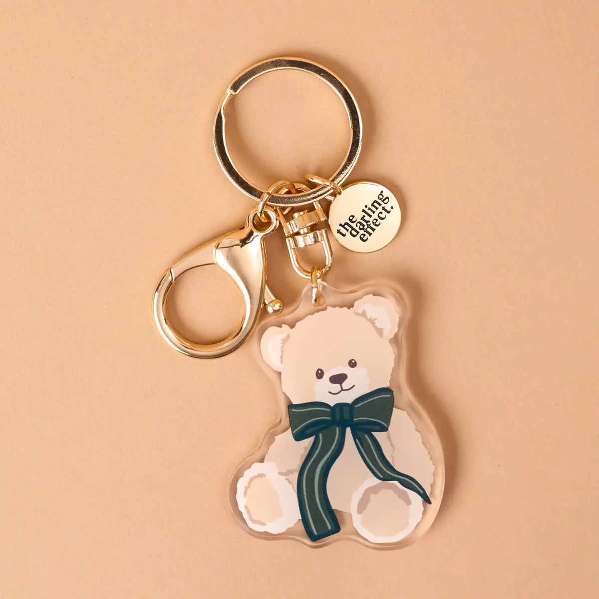 A clear acrylic keychain featuring an illustrated cream-colored teddy bear with a dark green bow, attached to gold-tone metal hardware including a key ring, lobster clasp, and a round branded tag that reads 'the thing direct'.