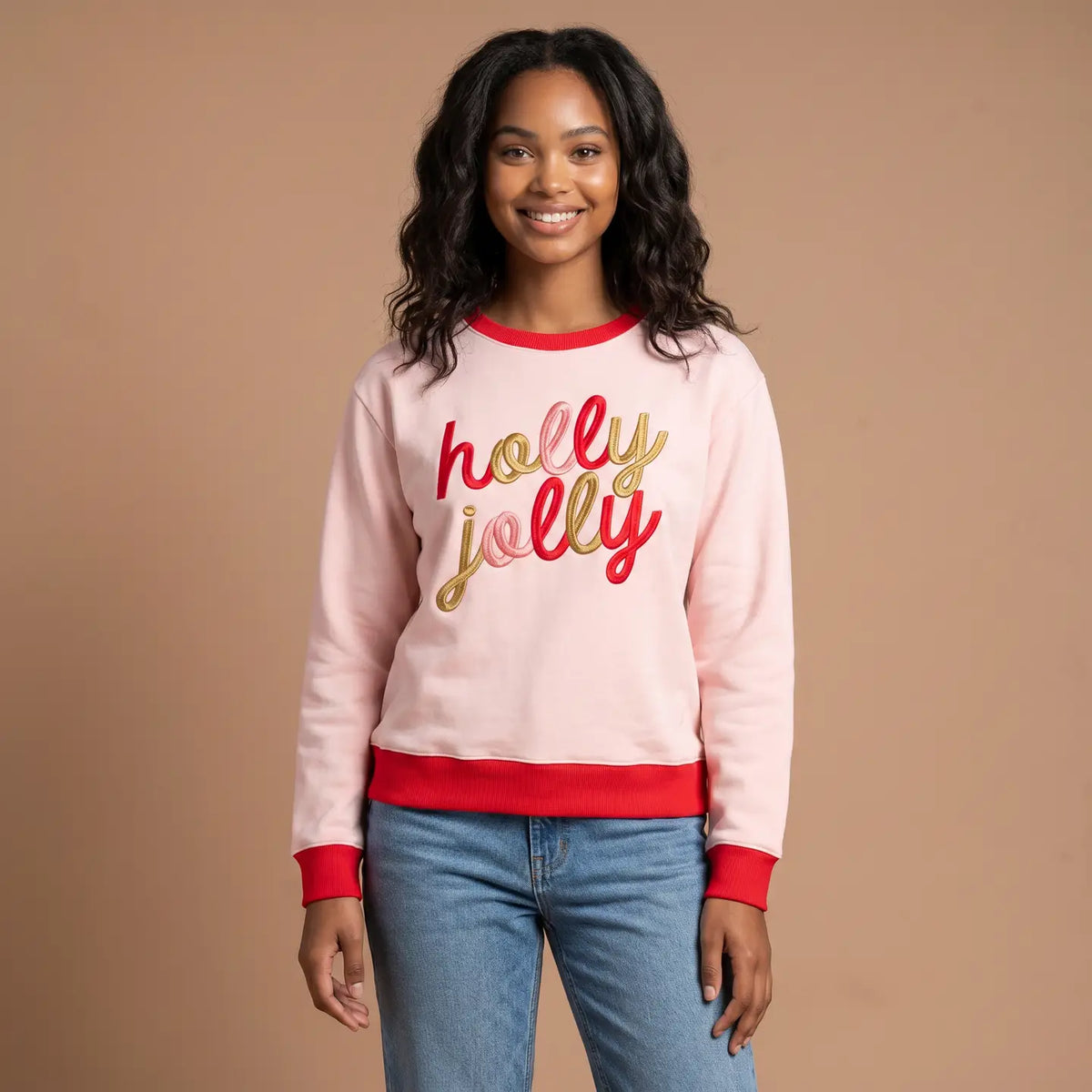 A woman wearing a light pink crewneck sweatshirt with red ribbed trim and 'holly jolly' text in red and gold across the chest, paired with blue jeans, against a solid tan background.