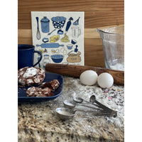 Kitchen counter with baking items including a plate of cookies, eggs, measuring spoons, and a dish towel with kitchen illustrations.