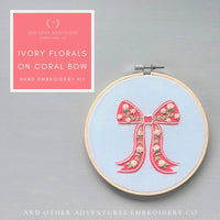 Embroidery hoop with floral bow design on a gray background, featuring 'And Other Adventures Embroidery Co' branding.