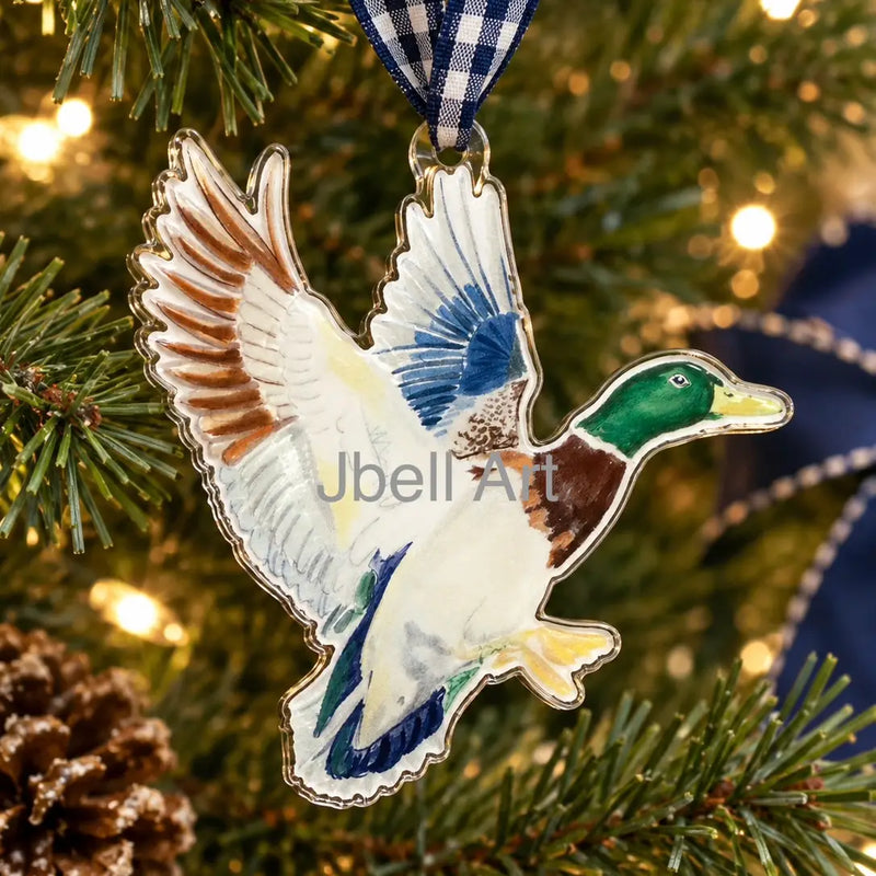 A flying mallard duck ornament hanging from a blue and white gingham ribbon on a Christmas tree with green pine branches and warm string lights in the background. The ornament has a white base with blue, brown, green, yellow, and black painted details.