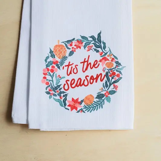 A white kitchen tea towel with a festive embroidered wreath design and the text 'tis the season' in red, displayed on a light wood surface.