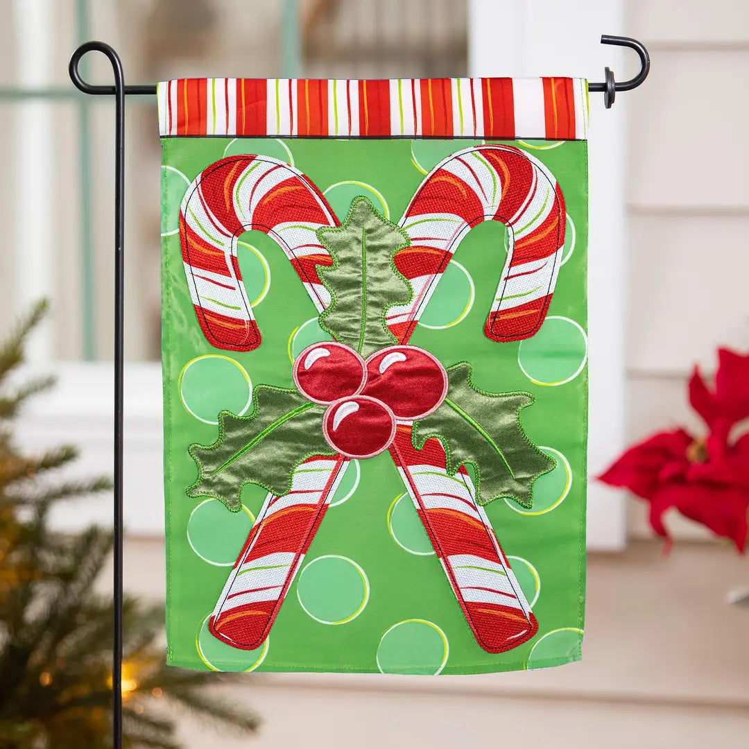 A vertical Christmas garden flag on a black metal stand, featuring two red and white striped candy canes, green holly leaves, and red berries on a bright green background with light green polka dots. The flag has a striped top band and is displayed outdoors in front of a house window.