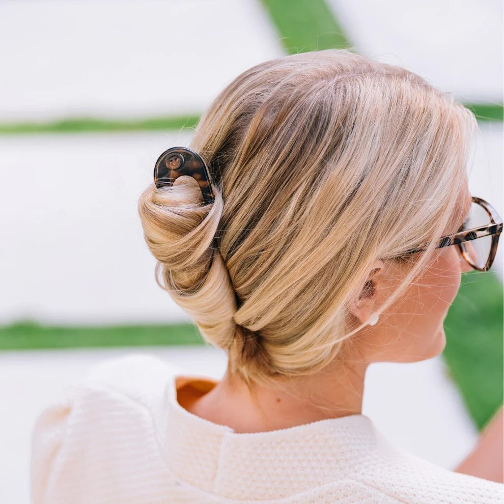 Woman with blonde hair styled in an updo, wearing glasses, with a blurred green and white background.