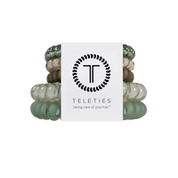 A set of six Teleties hair elastic bands in various shades of sage green, beige, and brown, bundled together with a white paper label featuring the black Teleties logo and brand name.