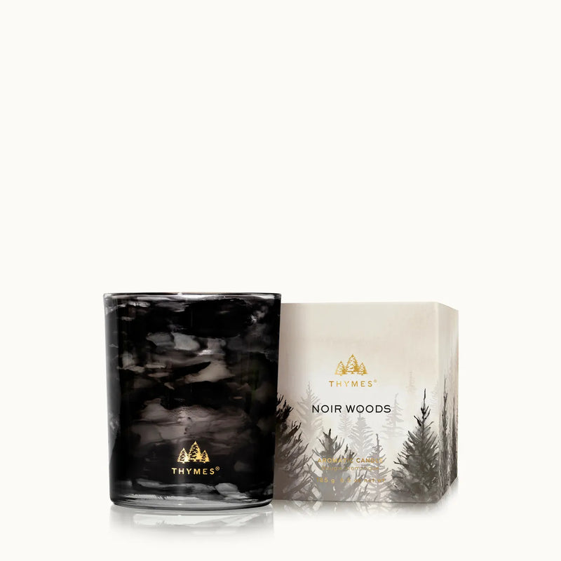 A black marbled scented candle in a clear glass vessel sits beside its illustrated packaging box on a clean white background. The box reads THÉMÉS and NOIR WOODS.