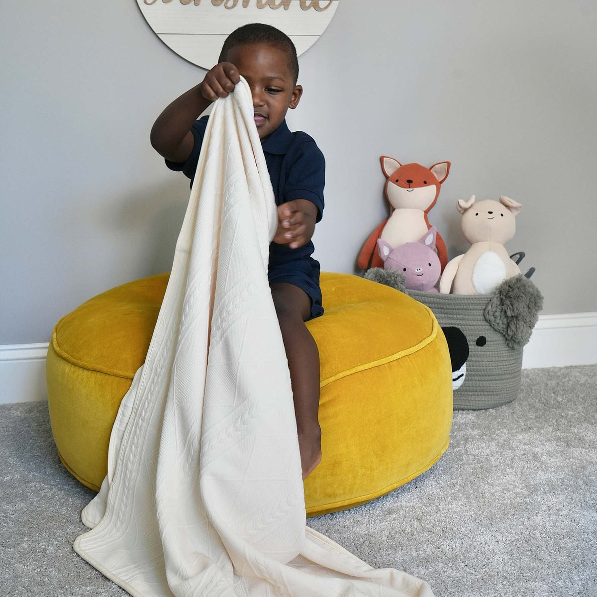 Child playing with a blanket on a yellow cushion in a room with stuffed animals.