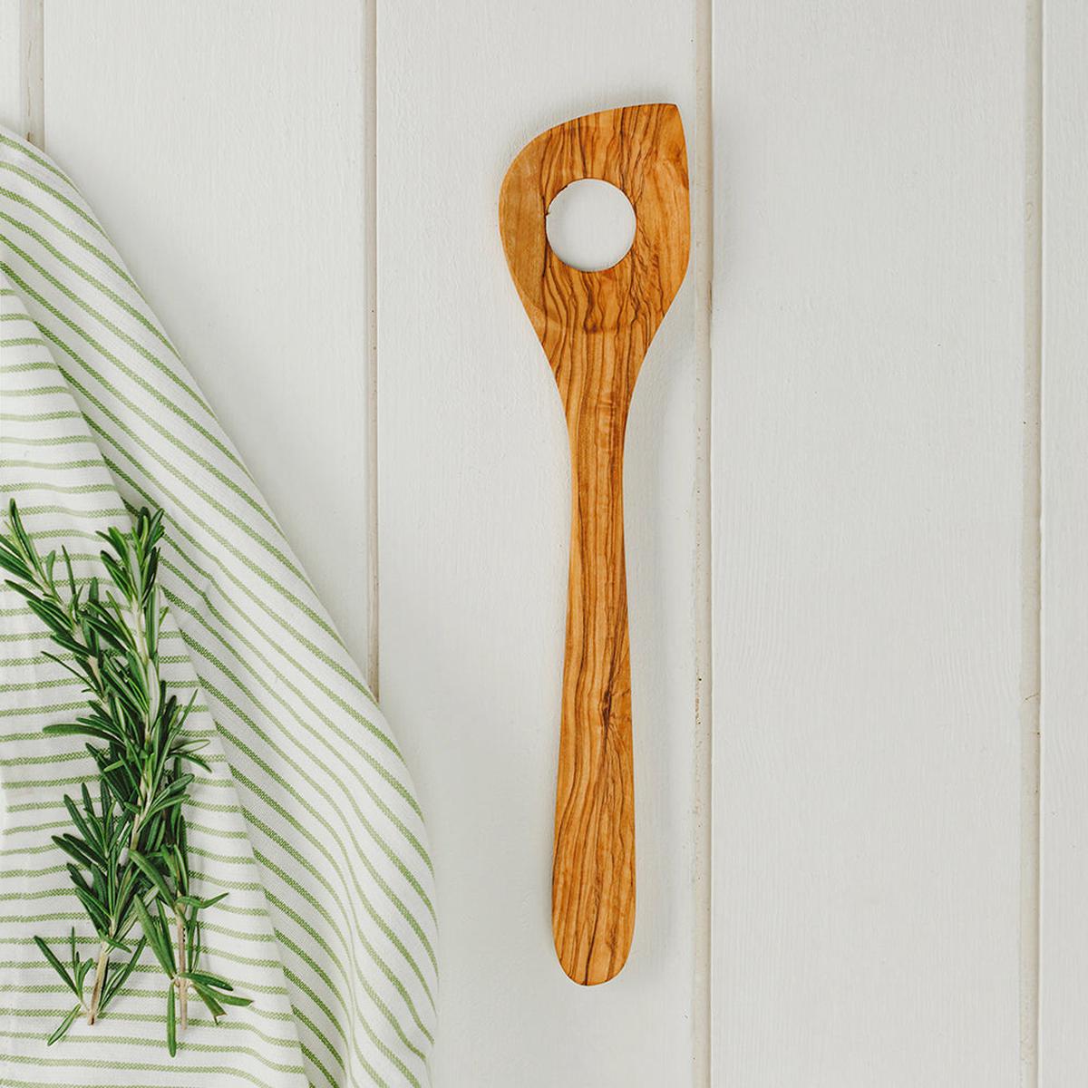 A wooden serving spoon with a circular cutout in the top, shown on a white wood plank background next to a green and white striped cloth and a sprig of rosemary.