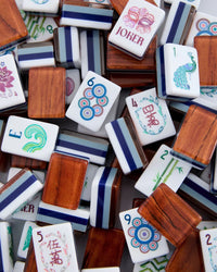 A pile of Mahjong tiles showing both the white backings with blue stripes and the wooden tops, including a visible 'JOKER' tile and numbered tiles with traditional Chinese and Western motifs.