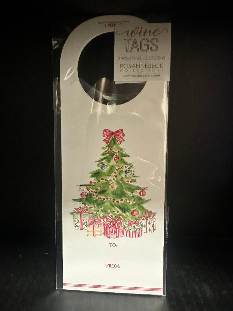 A packaged Christmas-themed wine tag from Rosanne Beck Collection, featuring a watercolor illustration of a green Christmas tree with ornaments, wrapped gifts, and a red bow on a white card, shown in clear plastic hanging packaging against a black background.