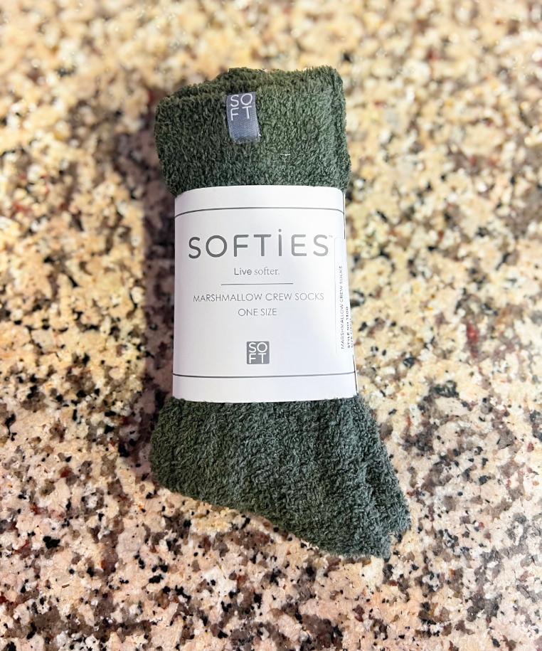A pair of dark olive green marshmallow crew socks rolled up with a white paper label that reads 'SOFTIES' and 'Live wool', placed on a speckled granite countertop.