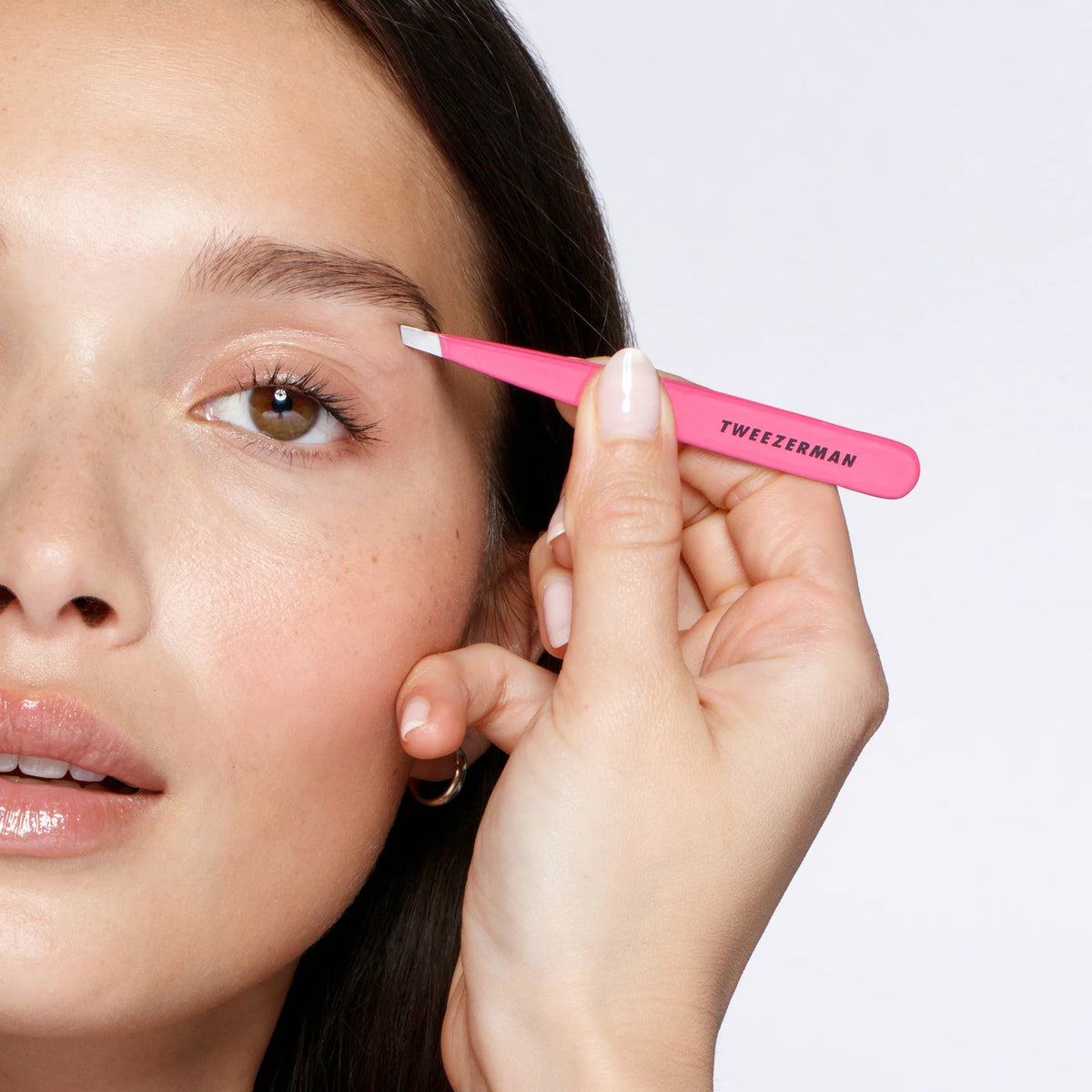 Person using a pink Tweezerman tool on their eyebrow against a white background